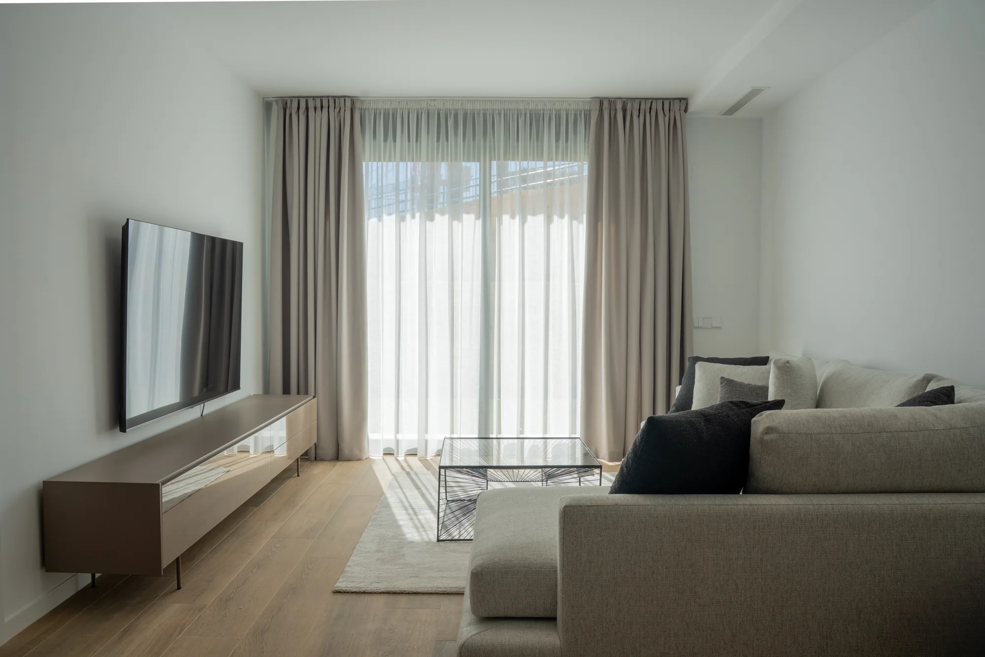 Salón con cortinas y visillo a medida en un ventanal de suelo a techo, luz natural sobre sofá gris